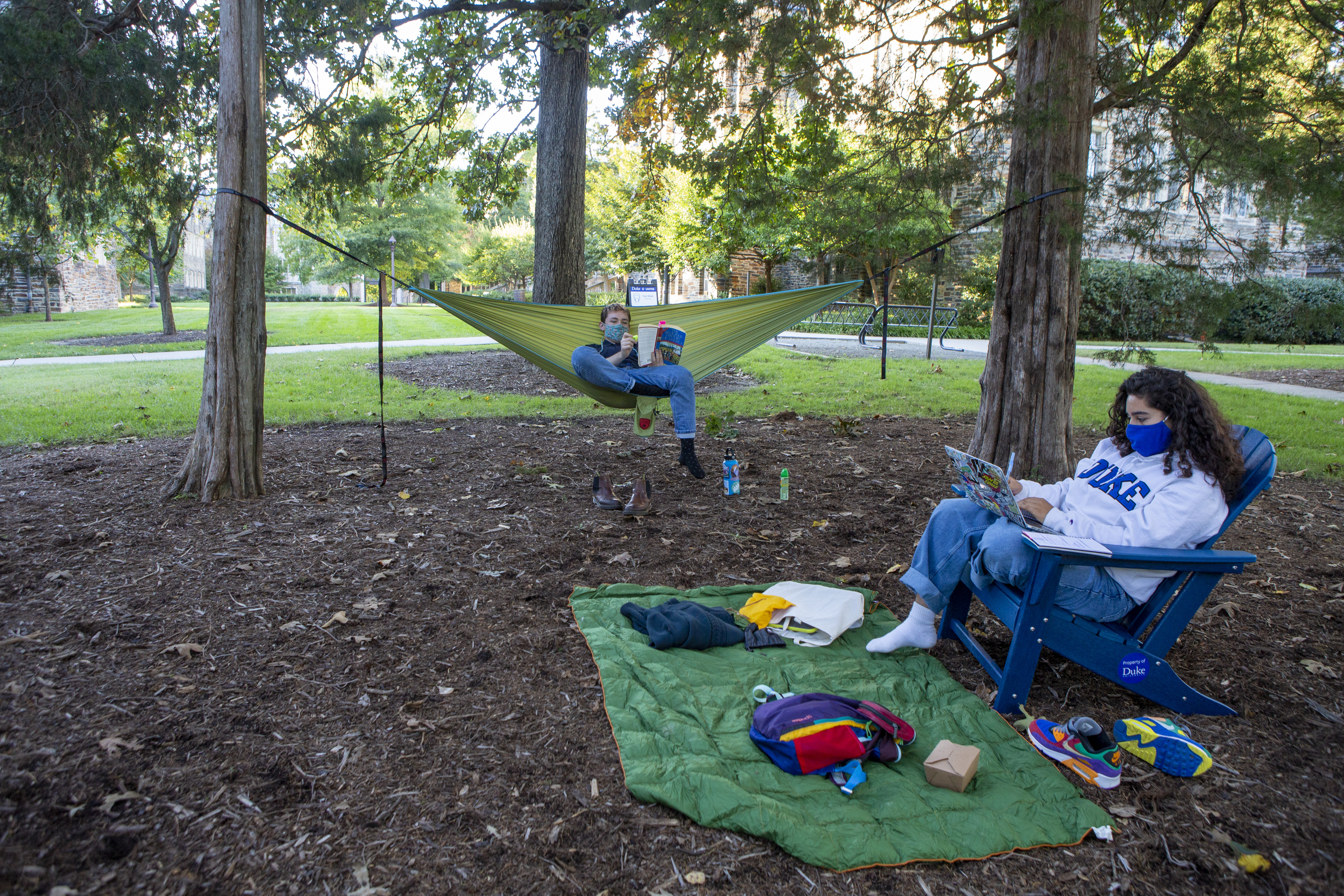 Two masked students study while masked and socially-distanced outside, one reading a book in a hammock and another typing on a laptop in a lawn chair.