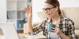 Woman waves at her computer screen holding coffee.