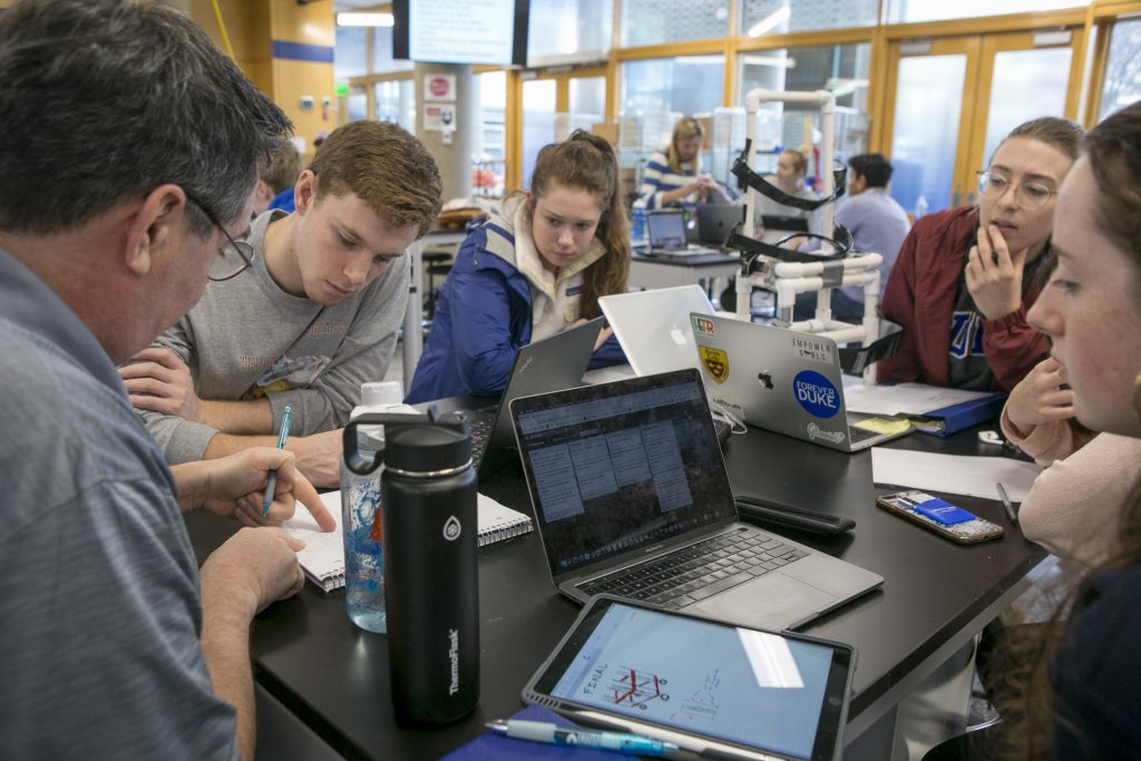 A professor explains something to a group of students using computers and an iPad.