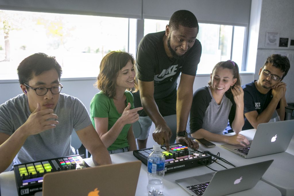 A student seated at a laptop connected to a soundboard explains something to her professor while neighboring students listen.