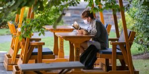 A student wearing a mask reads under green foliage.