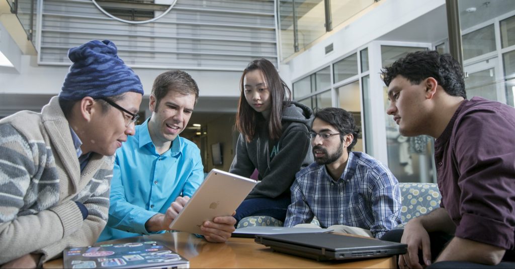 A group listens to a man gesturing to an iPad.