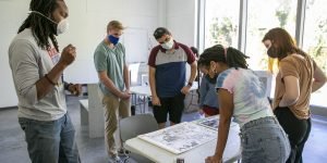 Students gathered around a table inspecting artwork while one student talks.